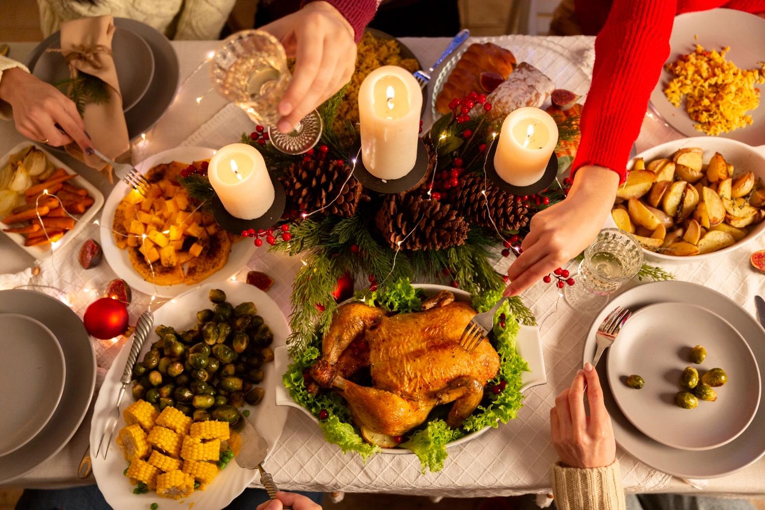A table filled with Christmas food including a roasted chicken, corn, brussel sprouts, carots, potatoes, candles and decorationsk, representing one of the best Chrsitmas traditions for couples: hosting the Christmas feast.