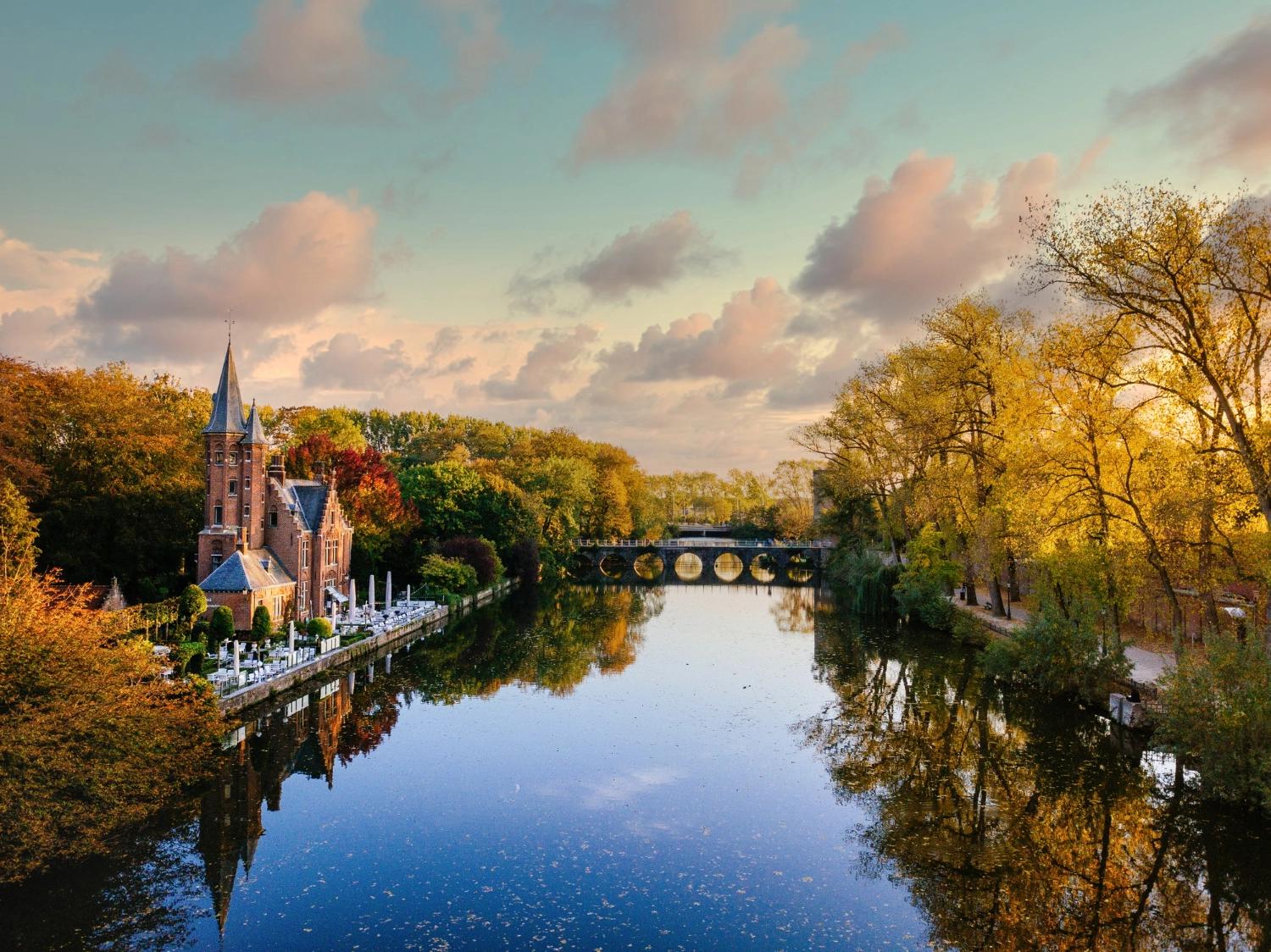 A view of a canal and castle in Bruges surrounded by colorful fall trees, capturing travel inspiration for romantic fall date ideas.
