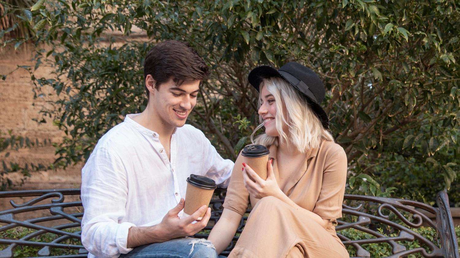 A man and a woman sitting on a bench and drinking coffee while answering some questions for couples to get to know each other better.