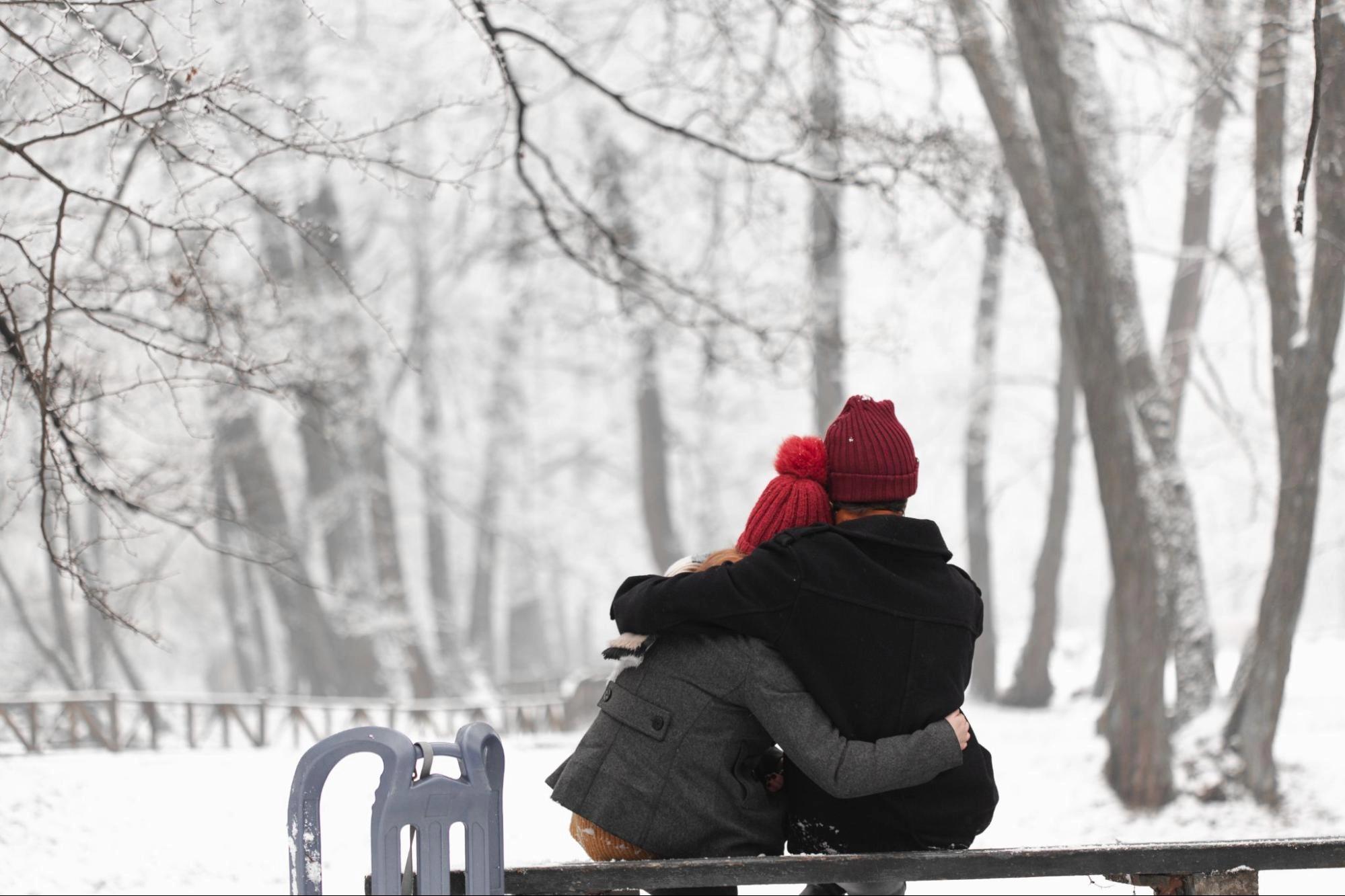 A couple sitting on a bench in a snowy park, holding each other, which is one of the best winter date activities for couples.