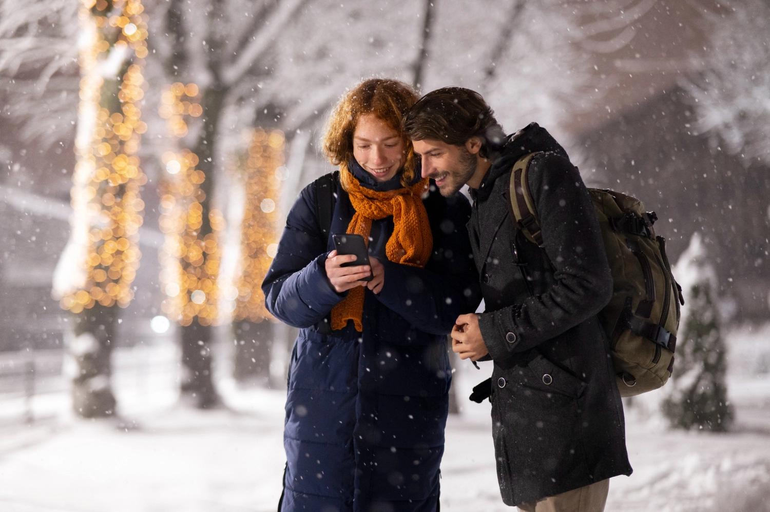 A couple looking at a phone while on weekend winter getaways in a snowy park, with snow-covered trees in the background.